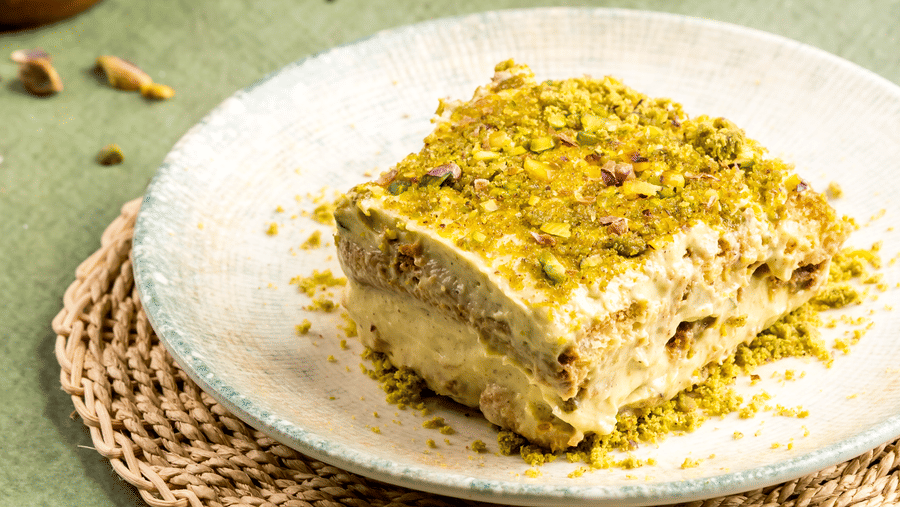 Slice of pistachio-topped layered dessert on a plate, with the remaining dessert in a glass dish in the background, served at Zaika in Gemini Continental.