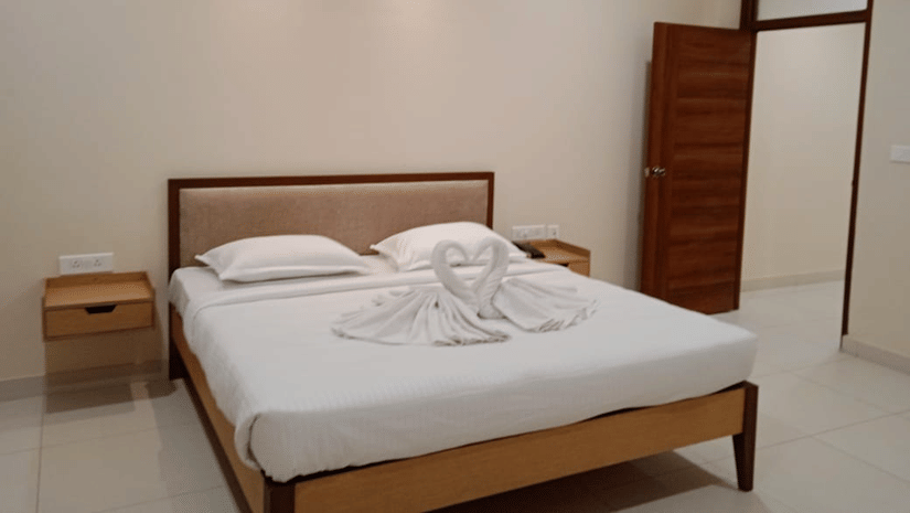 Bedroom at A M Suites with double bed, white bedding, swan-shaped towel sculptures forming heart, beige walls, tiled floor, wooden nightstands, open door, and recessed ceiling lighting.