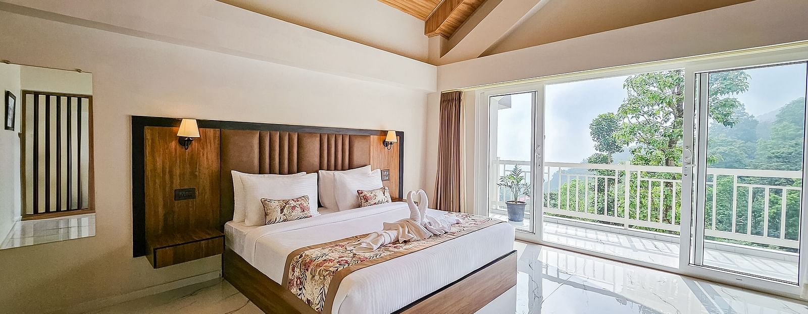 A luxurious bedroom with a large bed, wooden furniture, and a sliding glass door opening to a balcony with green views, at Yashshree Resorts & Spa, Kalimpong.