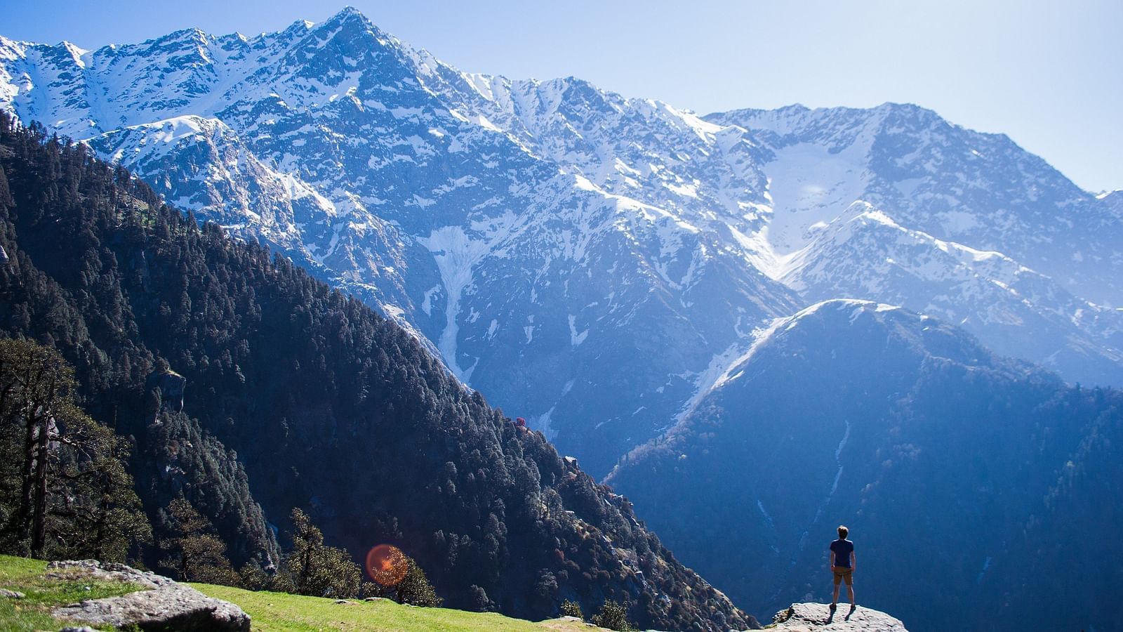 An image of snow caped mountains where a person is seen standing near a cliff enjoying the view