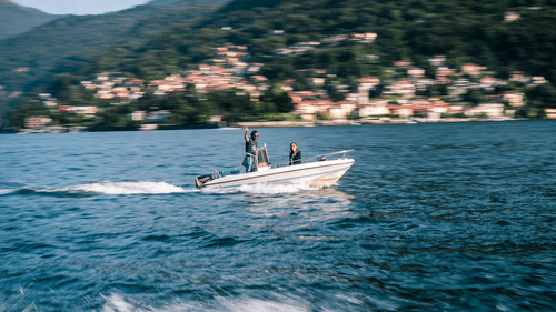 A small motorboat with 2 people on board moves across blue water, leaving a white wake, with a hillside town in the background