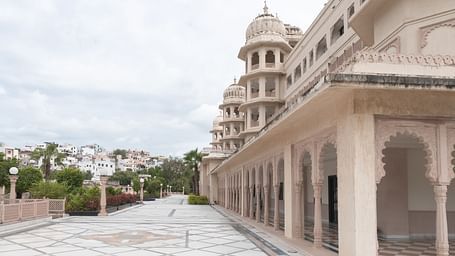 An elegant exterior view of Shri Vallabh Vilas Lords Plaza Nathdwara, showcasing intricately carved arches, domed pavilions, and a spacious marble courtyard lined with decorative lamp posts and greenery.