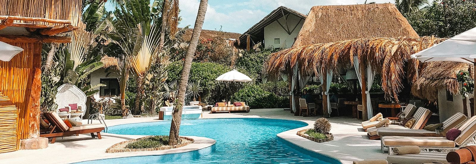 Lush tropical resort with a swimming pool, thatched-roof bungalows, and tall palm trees under a bright blue sky, suggesting a relaxing vacation.
