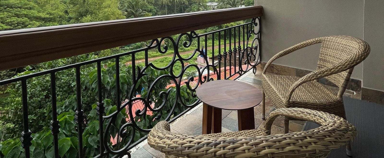 A serene balcony at The Evren, Vagator, showing two wicker chairs and a small table. The balcony overlooks a lush, green landscape with palm trees and fields under a cloudy sky.