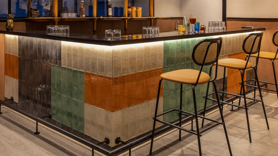 A bar counter inside Koi Garden restaurant. Bar stools stand in front of the counter structure. Shelves hold bottles and glassware beneath plants hanging from the ceiling.