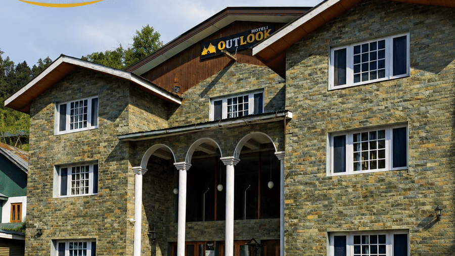 The exterior facade of Hotel Outlook, Pahalgam, showing a multi-storey stone building with windows, viewed from a lawn area with seating.