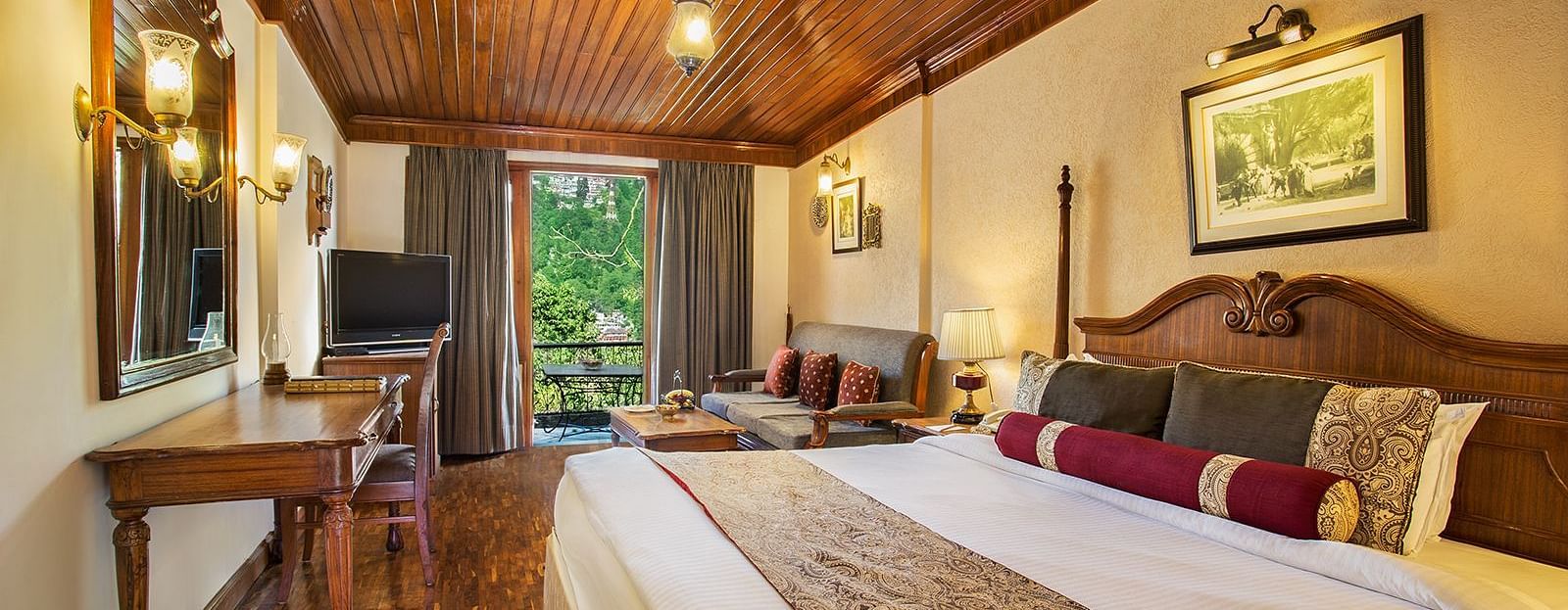 An image of a lake view room in naini retreat, nainital, with a king-size bed, wooden furniture, and a bay window overlooking a garden. The room features a ceiling fan, a television, and a small sitting area and wooden flooring.
