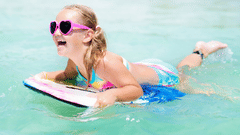 A young, smiling girl wearing pigtails and heart-shaped sunglasses floating over water on a body board.