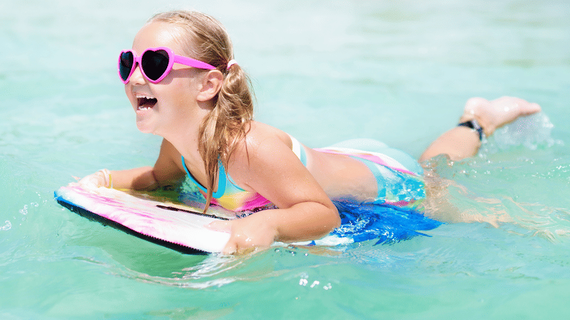 A young, smiling girl wearing pigtails and heart-shaped sunglasses floating over water on a body board.