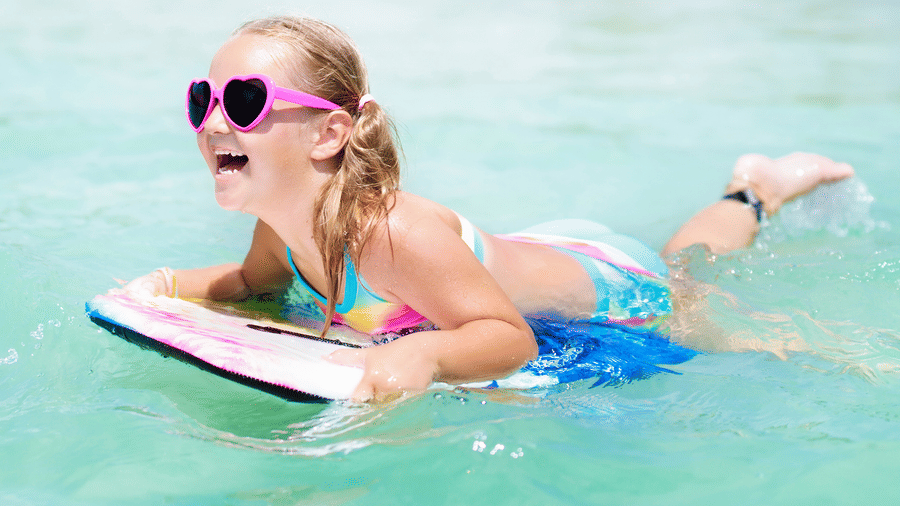 A young, smiling girl wearing pigtails and heart-shaped sunglasses floating over water on a body board.