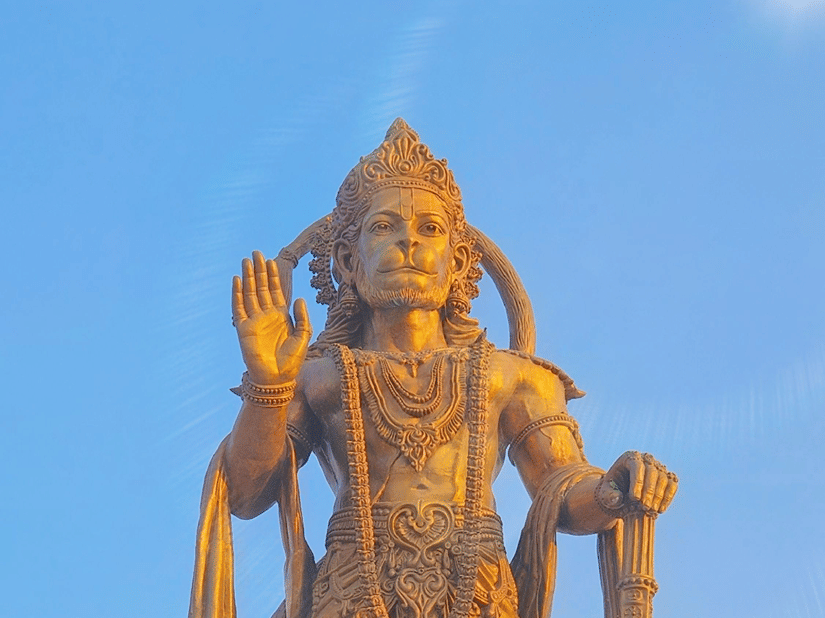 A tall golden statue of a Hindu deity standing on a decorated pedestal against a clear blue sky.