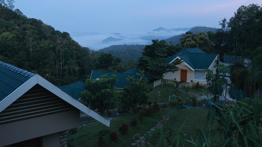 A dimly lit facade of Aveda Mountains and Mist Resort and Spa, Munnar, featuring the villas and the lush green surroundings during early morning hours.