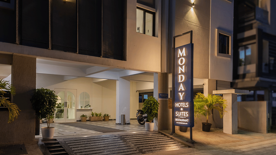 Lit vertical hotel sign beside the entrance steps, potted palms and the covered porch with glass doors at night at Monday Hotels & Suites Madhapur, Hyderabad.