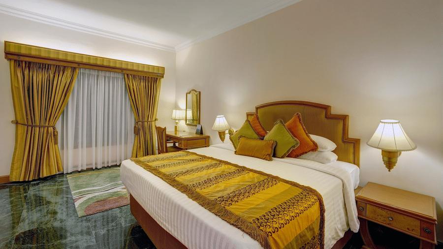 Royal Suite with king size bed, golden accents, bedside lamps, classic furnishings, and floor-to-ceiling curtains overlooking a private balcony at The Ummed Ahmedabad