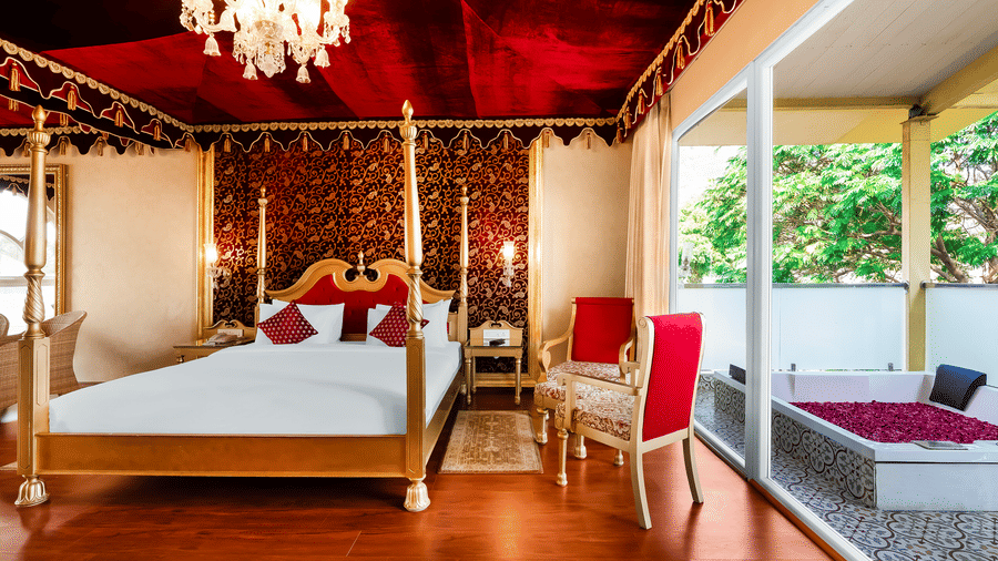 A room at Rajasthali Resort & Spa with a canopy four-poster bed, ornate decor and rich red furnishings, opening onto a balcony with a bathtub, offering a luxurious stay in Jaipur.