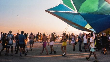 A colourful abstract sculpture with people standing and dancing around it in a desert at sunset.