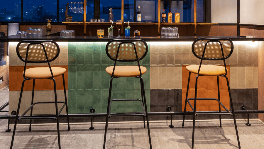 View of the bar area at Koi Garden. Three bar stools face the serving counter. Shelves with bottles and decorative items sit behind the bar beneath hanging greenery.