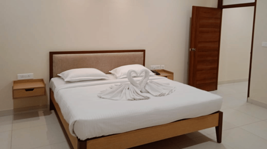 Bedroom at A M Suites with double bed, white bedding, swan-shaped towel sculptures forming heart, beige walls, tiled floor, wooden nightstands, open door, and recessed ceiling lighting.