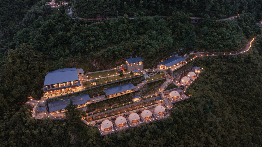 Aerial night view of a luxury hillside resort featuring illuminated dome cabins surrounded by lush forest.