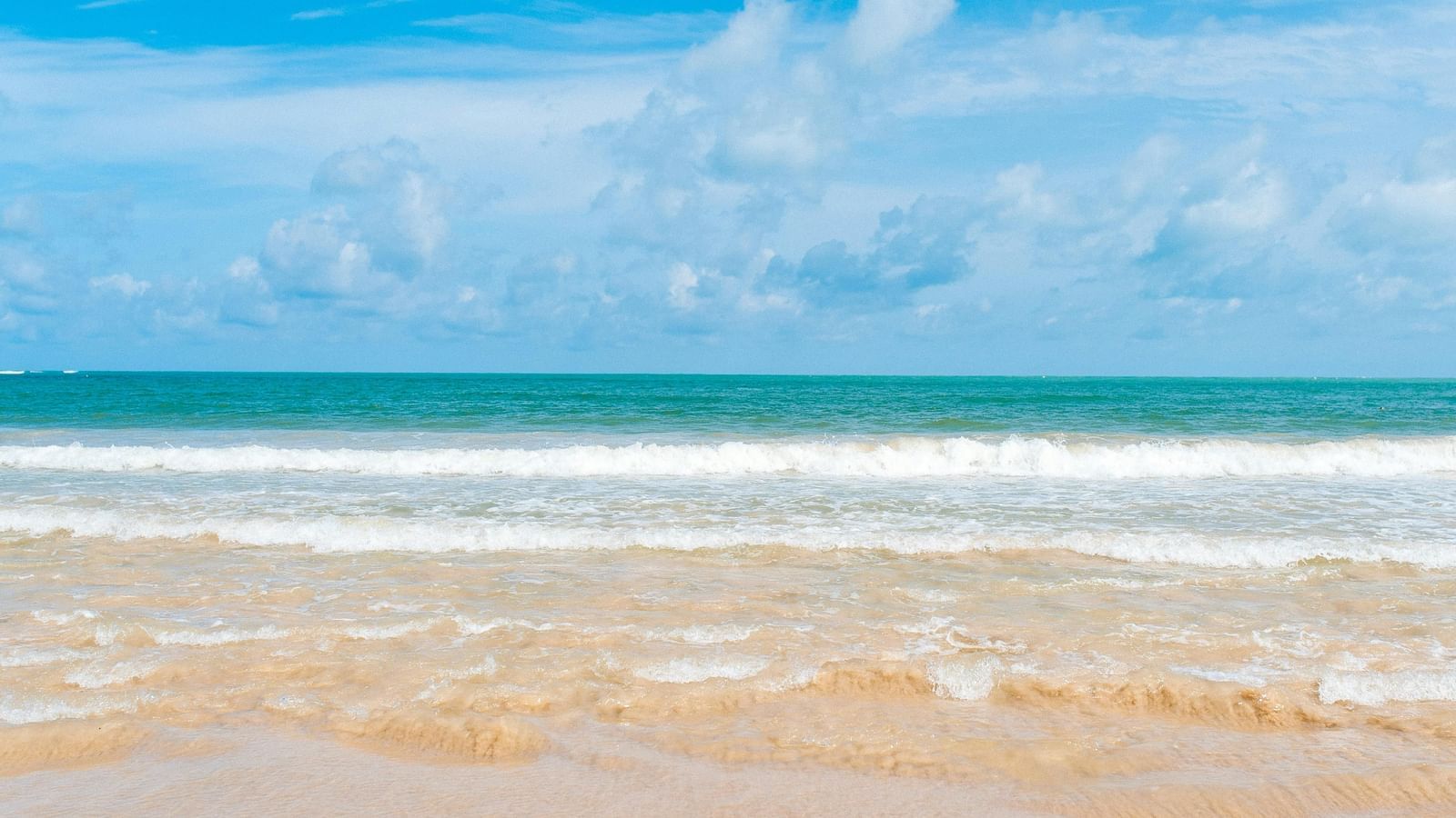 A calm, sunlit beach with soft golden sand and gentle waves rolling in from the turquoise sea under a cloudy blue sky.