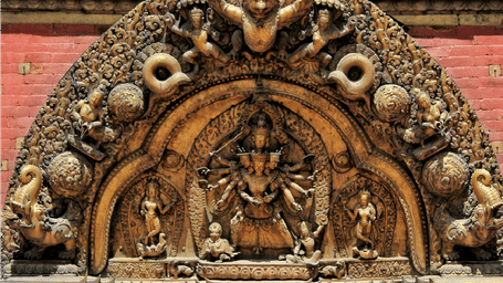 Intricately carved wooden torana archway depicting a multi-armed deity central figure topped with a winged Garuda motif above an entrance.