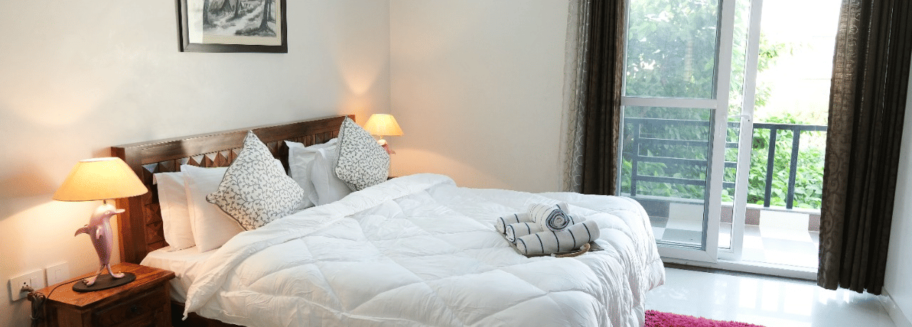 An image of a bedroom featuring a bed, a bedside table with lamp, anda a glass door leading to the balcony - LIme Tree 3BHK Serviced Apartments Near Medanta HospitalAn image of a bedroom featuring a bed, a bedside table with lamp, anda a glass door leading