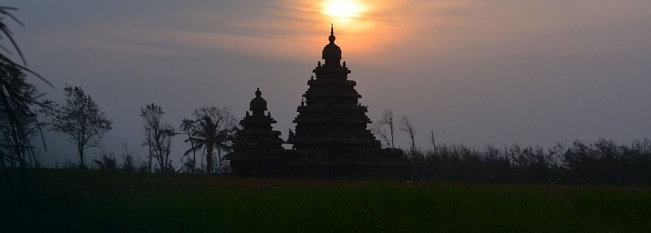 silhouette of a architectural figure with two pointed roofs in a setting of trees with the sun in the distance 