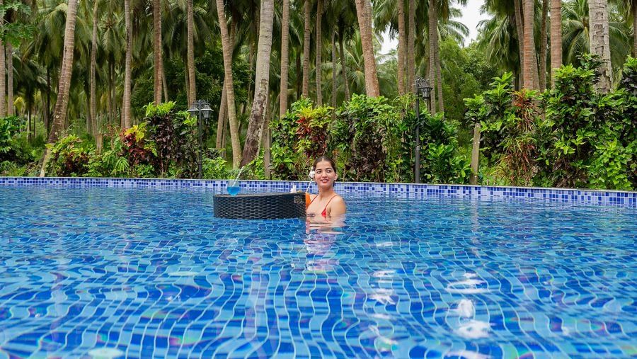 Woman enjoying a drink at a floating tray in a swimming pool surrounded by a dense, tall coconut palm forest.