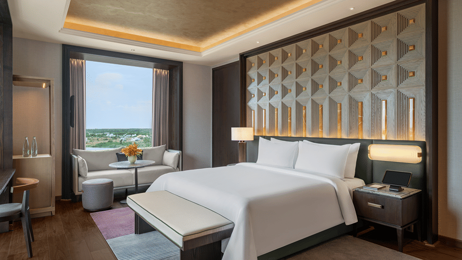 Presidential Suite second bedroom with a double bed, padded bench, a sofa by the window and a carved headboard wall at The Residency Signature Madurai.