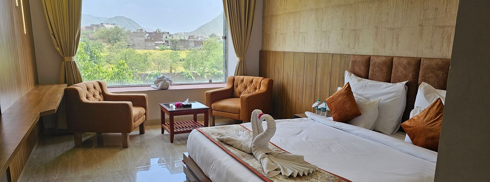 A bright and spacious resort room at Bamboo Saa Mulberry Resort, Pushkar, featuring a large window with a view of the outdoors including a made bed with a patterned runner, two armchairs, and a small table.