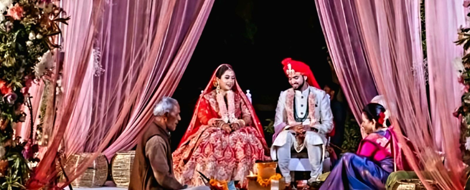 A couple sits under a beautifully decorated canopy with floral arrangements and drapes during an outdoor ceremony
