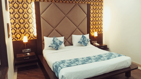 A king size bed with a padded headboard, a patterned wallpaper background, 2 bedside tables, and 2 illuminated wall lamps at Hotel Mumbai House, Thane.