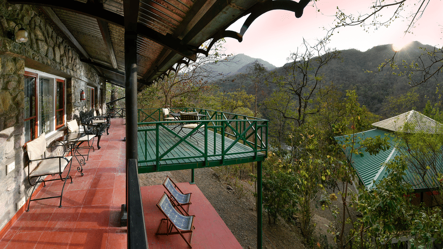 Wide angle view of a long veranda with a red floor and green wooden railings looking out over a lush green valley and mountains at Neemrana’s Glasshouse on The Ganges offering a peaceful outdoor space for guests to relax and soak in the scenery