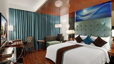 A spacious deluxe hotel room with a large double bed, decorative cushions, wooden flooring, a padded headboard, a wooden desk with chair, soft lighting, and curtains