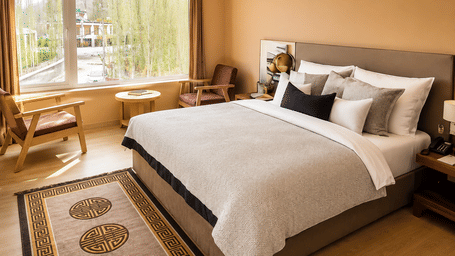 Hotel room with a bed dressed in white linen, a wooden headboard, a patterned rug and a large window overlooking mountain views close by.