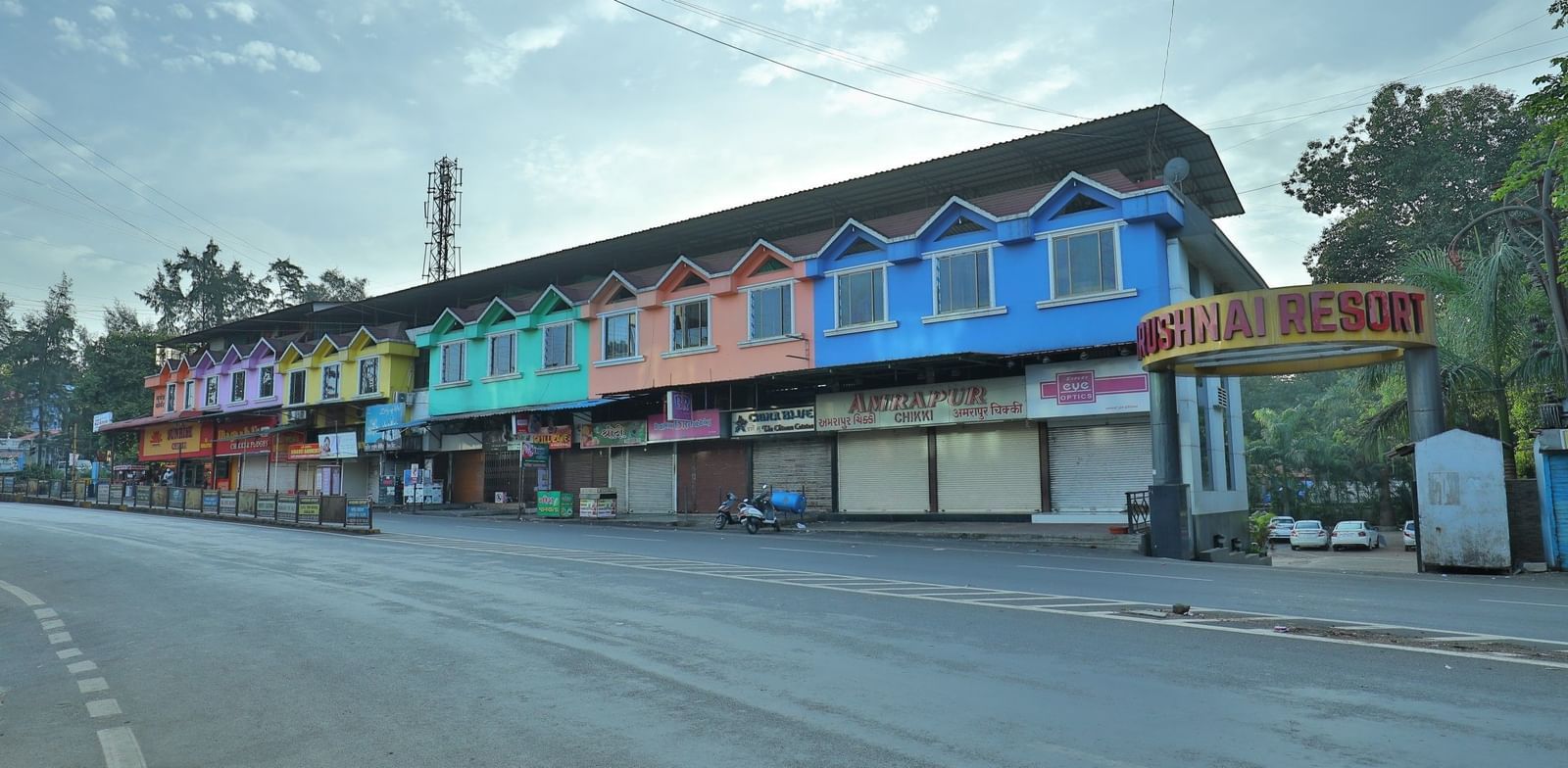 The Krushnai Resort sign marks a long building with colourful facade lining a quiet road under a cloudy sky.