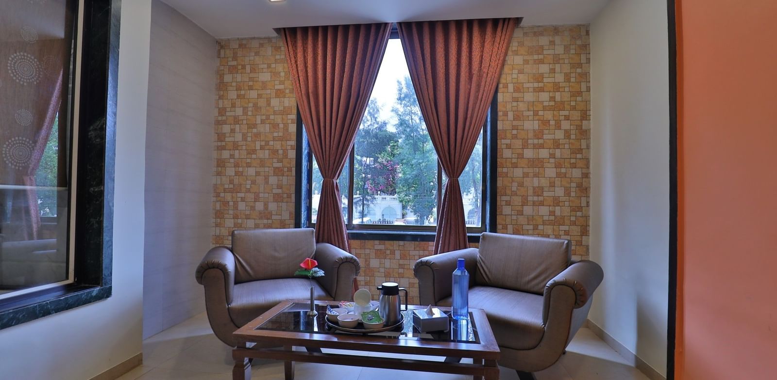 A seating area in a suite at Krushnai Resort with 2 lounge chairs, coffee table, and red drapes, beside a window.