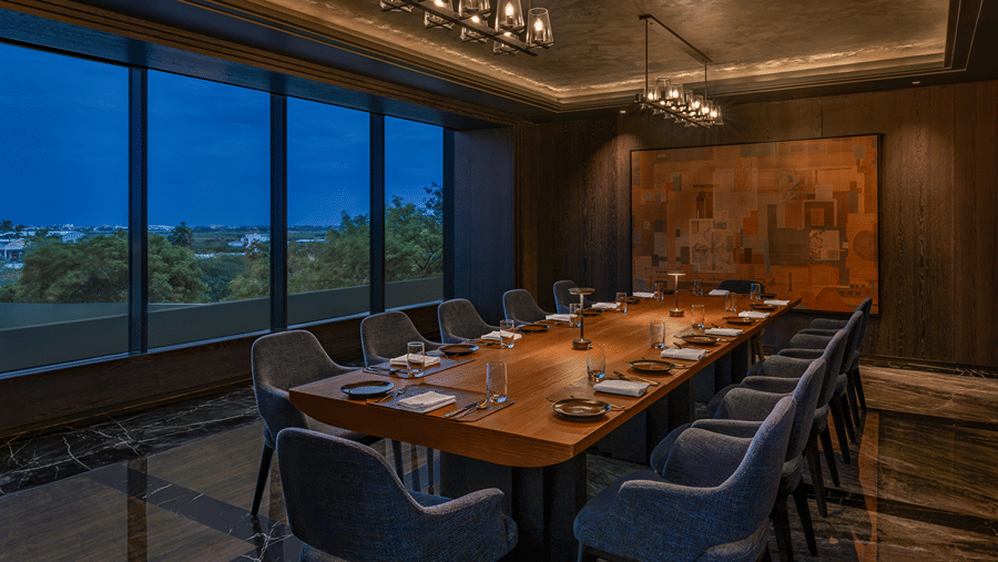 The Grand Trunk private dining room with a long wooden table, grey upholstered chairs, glass pendant lights and windows at The Residency Signature Madurai.