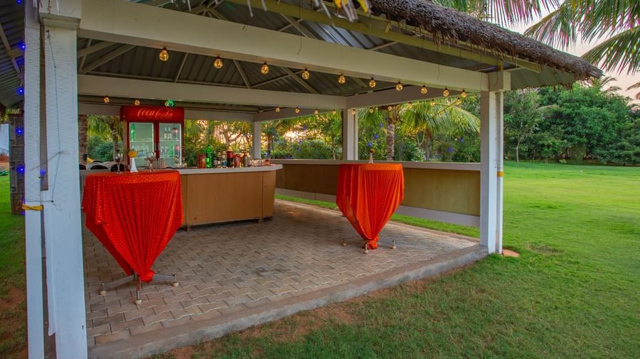 A charming outdoor bar or serving area with a thatched roof, red bar stools, and a wooden counter, set on a grassy lawn | Citrus Garden | Grand Bay Resort and Spa