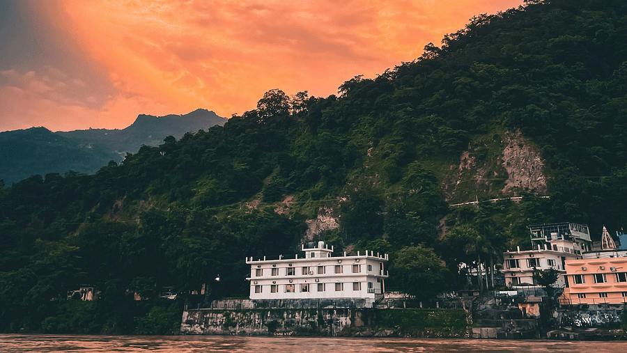 Rishikesh, Uttarakhand is captured at sunset with the Ganges River in the foreground and buildings nestled against a forested hillside under a colourful sky.