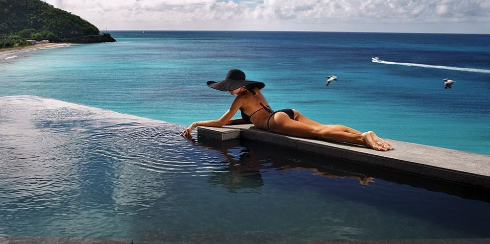 Woman lounging by a sea overlooking the turquoise Caribbean Sea, wearing a black bikini and wide-brimmed sun hat, with speedboats and birds in the distance.
