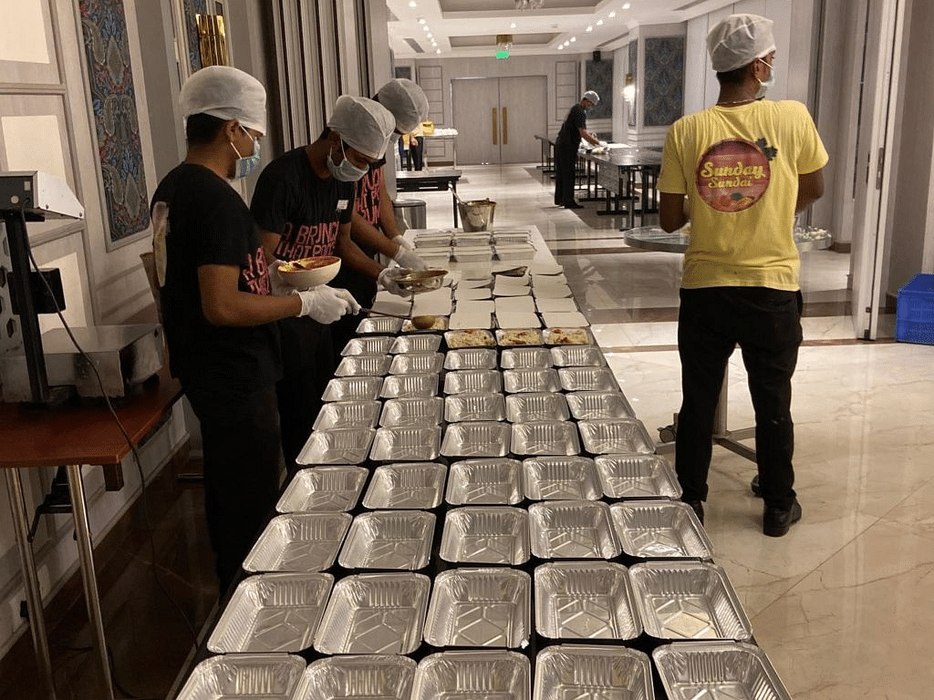 The staff filling up a lot of aluminium  foil containers with food wearing masks and gloves.