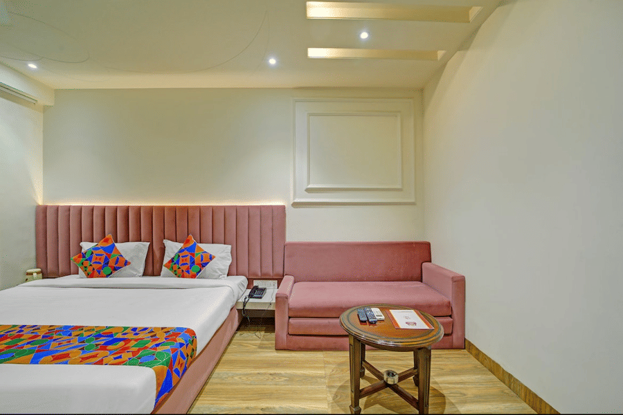 Double bed with patterned runner, pink headboard wall, small seating area and wooden flooring at Hotel Maurya, Bhopal.
