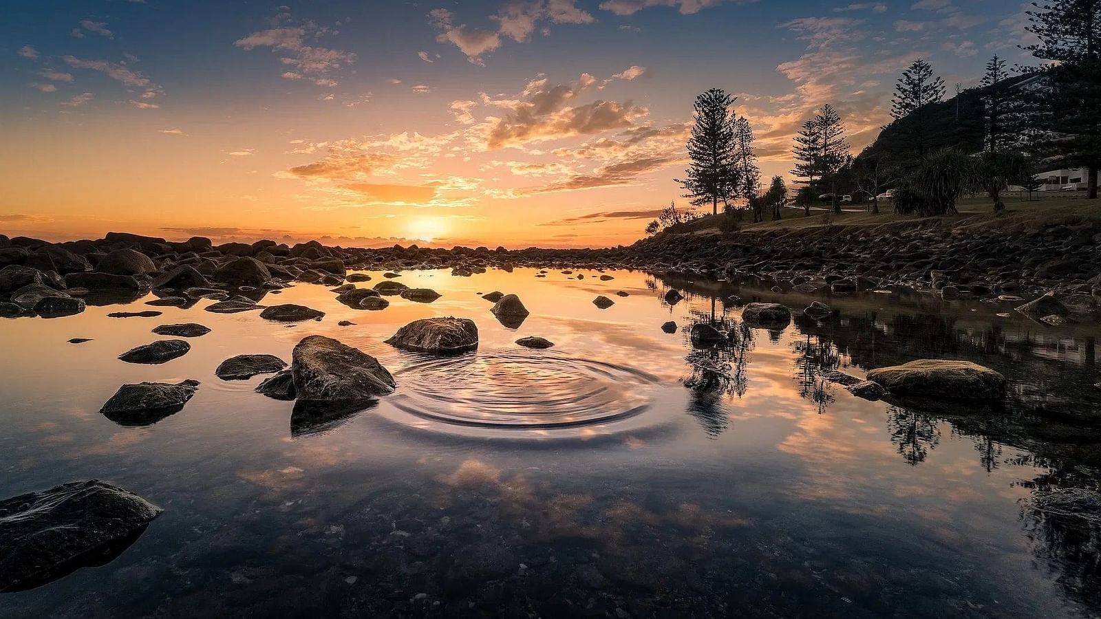 Sunset over a rocky shore, reflection in water, lighthouse visible.