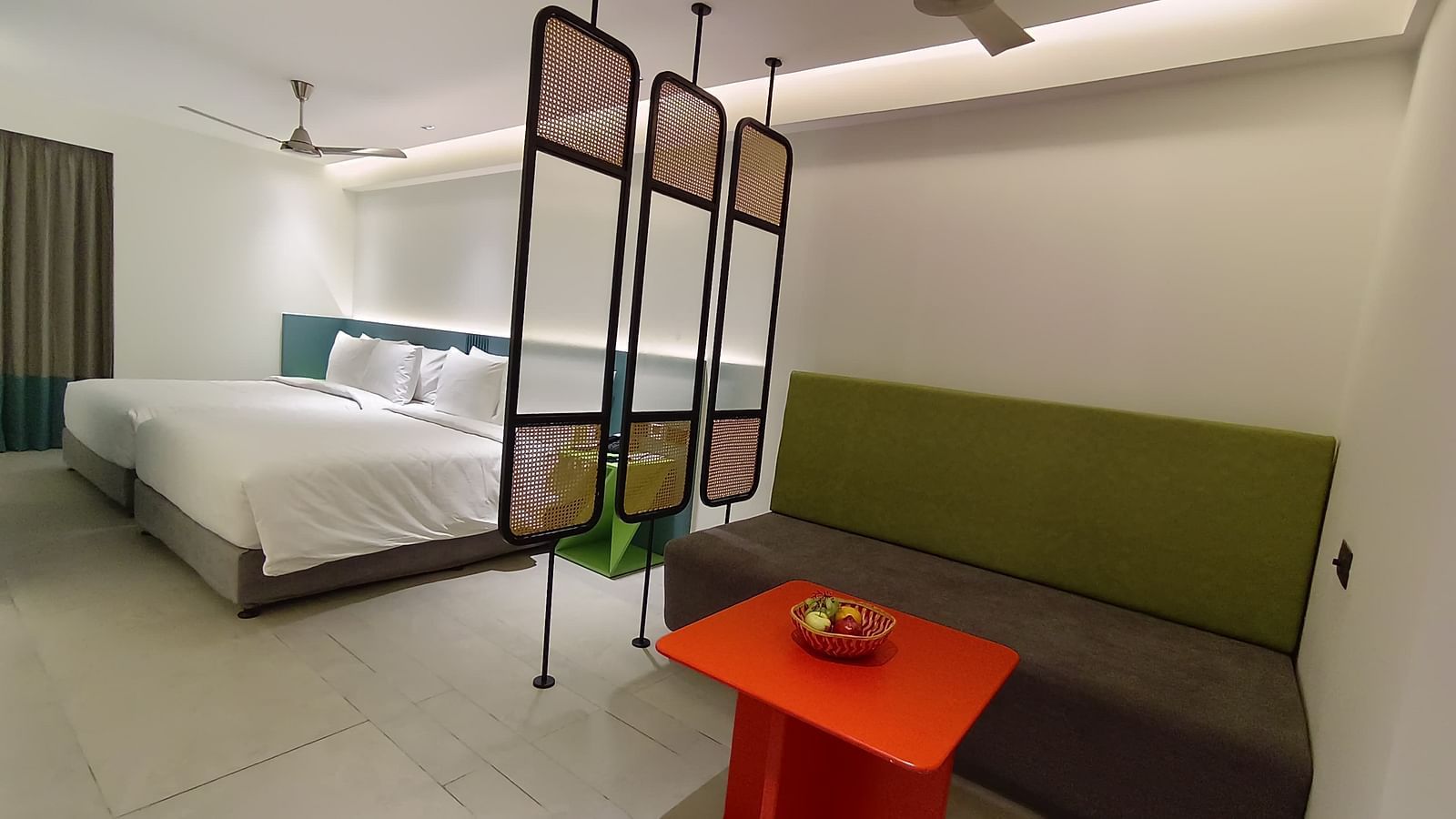 A spacious suite room featuring a neatly made bed with two double beds placed together, a separate seating area with a sofa and coffee table, and a partial divider between the sleeping and living spaces