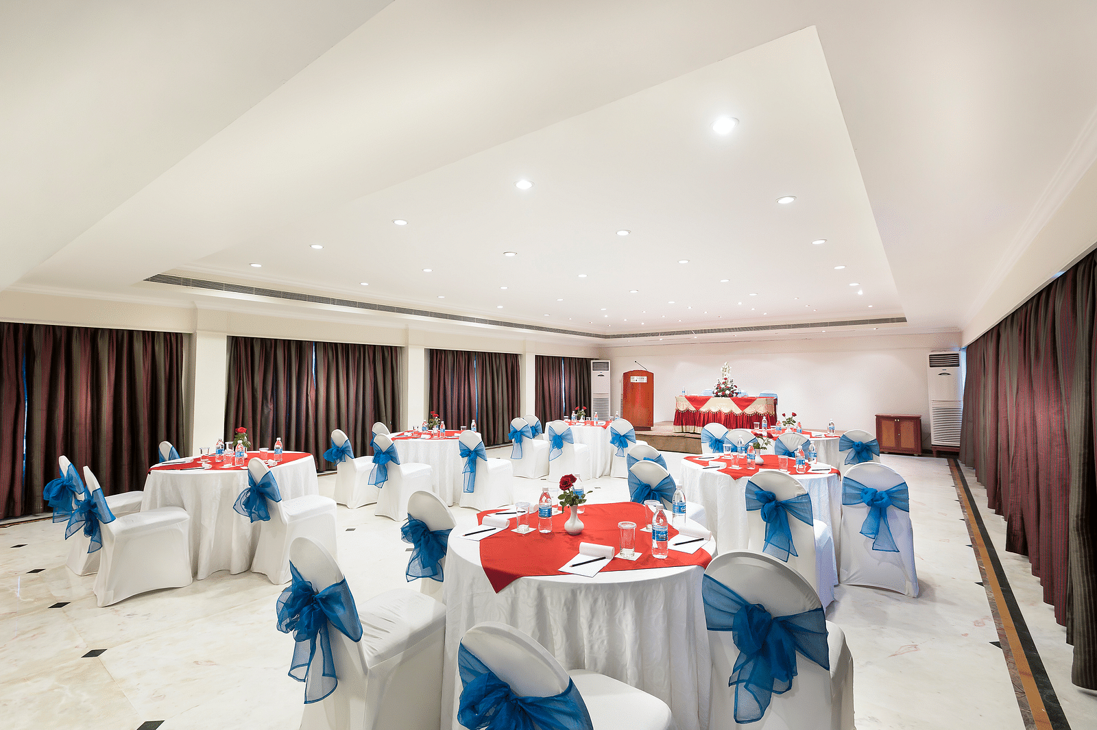 A banquet hall with white chairs laced with blue ribbons pointing towards the stage at Regency Kanchipuram by GRT Hotels.