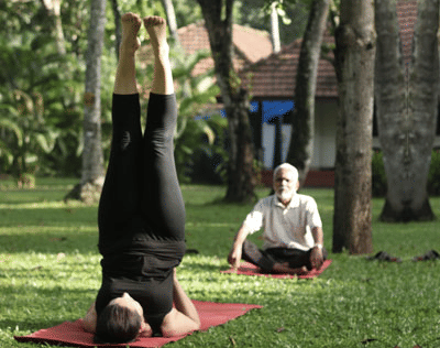 A woman performs a headstand (Sirsasana) on a red yoga mat in a sunny, grassy garden while an elderly man sits in meditation nearby.
