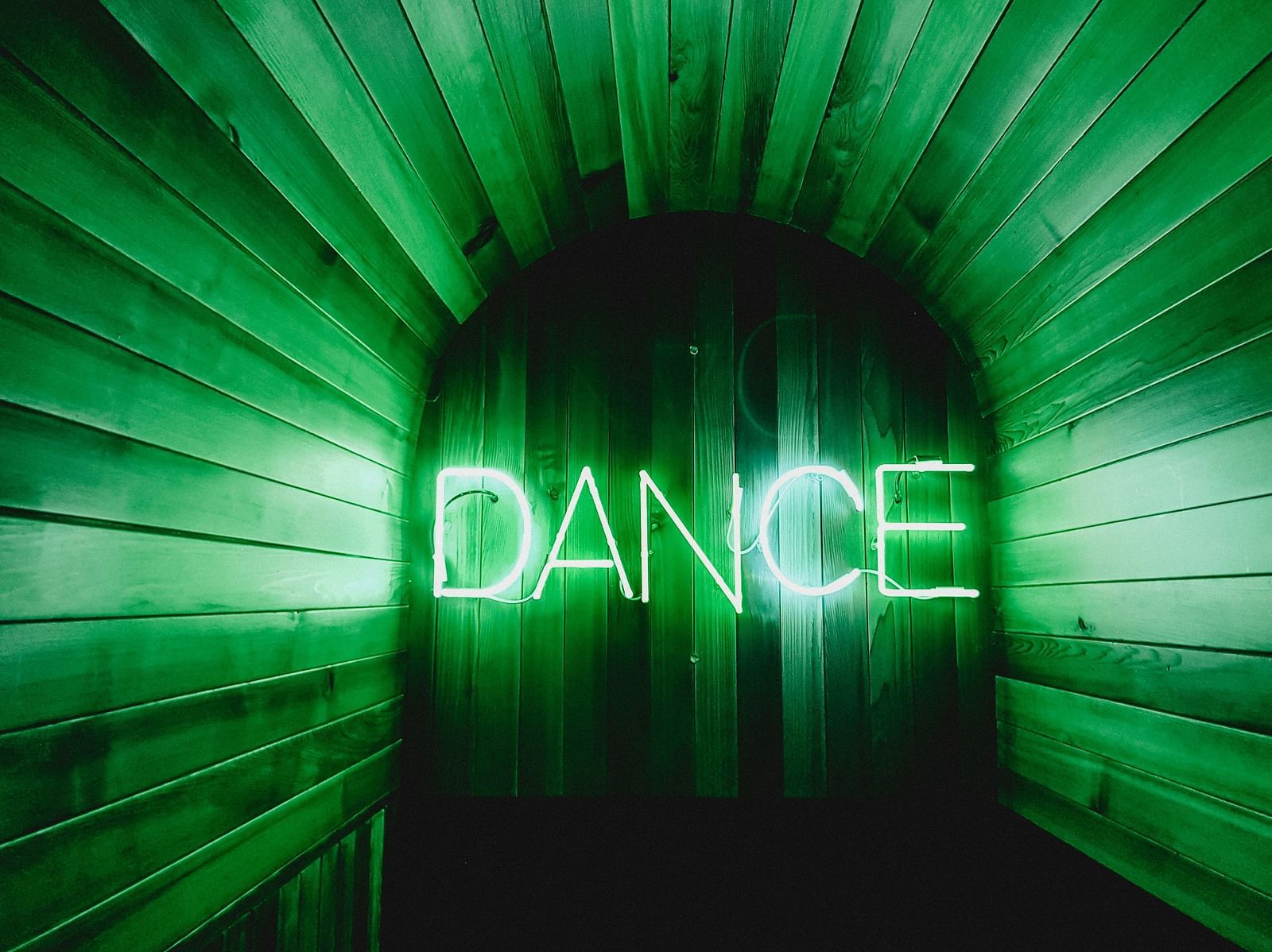 Dance written in green Neon lights with the light reflecting on the wooden walls