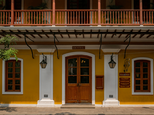 A symmetrical yellow facade featuring a central wooden door, white columns, lantern-style lights, and a balcony with a wooden railing above.