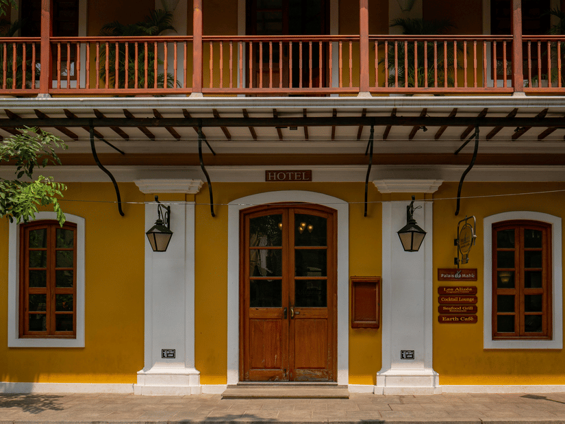A symmetrical yellow facade featuring a central wooden door, white columns, lantern-style lights, and a balcony with a wooden railing above.